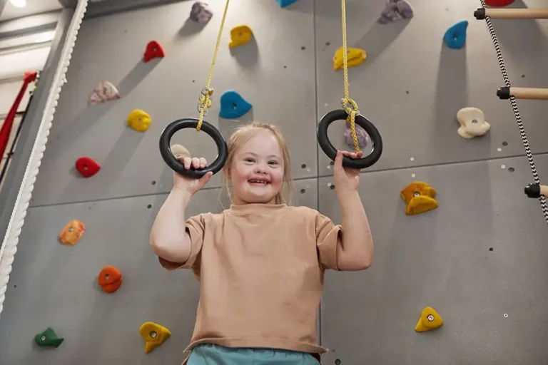 Waist up portrait of cute girl with down syndrome enjoying exercise at home or at sports center and looking at camera while holding at rings against climbing wall, copy space